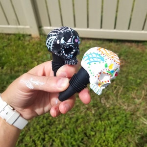 May include: Two decorative skull-shaped bottle stoppers. One is black with white and light blue accents, the other is white with colorful floral designs. Both have black rubber bases and are held in a hand against a green background.