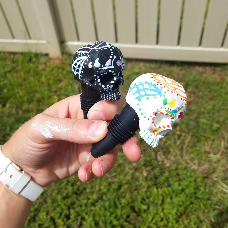 May include: Two decorative skull-shaped bottle stoppers. One is black with white and light blue accents, the other is white with colorful floral designs. Both have black rubber bases and are held in a hand against a green background.