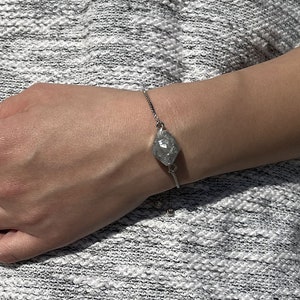 May include: A silver bracelet with a small, oval-shaped charm. The charm has a textured, metallic finish.