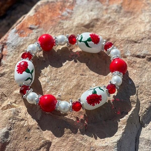 May include: A red and white beaded bracelet with red flowers painted on the white beads. The bracelet is on a rock.