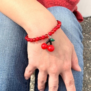 May include: A red beaded bracelet with a cherry charm. The bracelet is on a person's wrist.