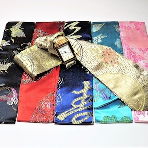 May include: Six different fabric watch bands, each with a unique design. The bands are in various colors, including black, red, blue, teal, and pink. The bands are made of a silky material with intricate patterns. A gold watch is placed on top of the bands.