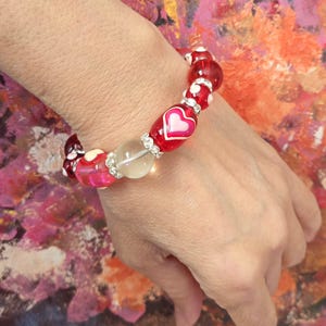 Puede incluir: Una pulsera de cuentas con cuentas rojas, rosas y transparentes. La pulsera incluye cuentas con dise&ntilde;os de coraz&oacute;n blanco y una cuenta roja grande con un coraz&oacute;n rosa. Los detalles de pedrer&iacute;a a&ntilde;aden brillo.
