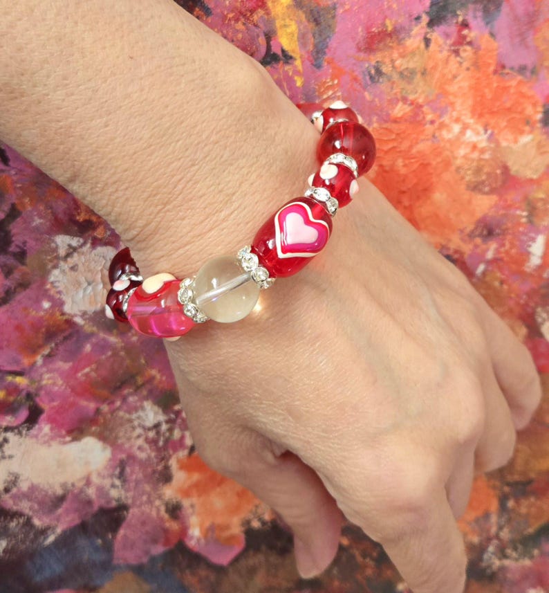 Puede incluir: Una pulsera de cuentas con cuentas rojas, rosas y transparentes. La pulsera incluye cuentas con dise&ntilde;os de coraz&oacute;n blanco y una cuenta roja grande con un coraz&oacute;n rosa. Los detalles de pedrer&iacute;a a&ntilde;aden brillo.