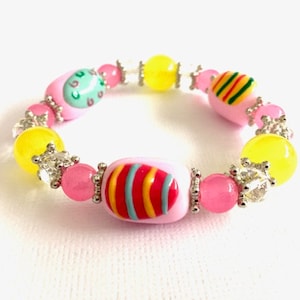 May include: A pink, yellow, and green beaded bracelet with colorful egg charms. The bracelet is made with small, round beads and features two egg charms with striped patterns and one egg charm with a green and white pattern.