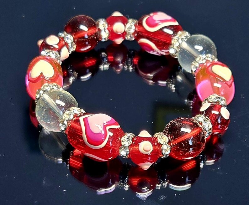 Puede incluir: Un brazalete de cuentas con cuentas rojas, transparentes y rosas. Algunas cuentas tienen dise&ntilde;os de coraz&oacute;n. El brazalete est&aacute; acentuado con espaciadores plateados. Las cuentas tienen aproximadamente 2 cm de di&aacute;metro.