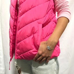 May include: A pink puffer vest with a zip fastening and a pocket on the left side. The vest is worn over a long-sleeved pink shirt and light blue jeans. The person is wearing a silver bracelet with a blue butterfly charm.