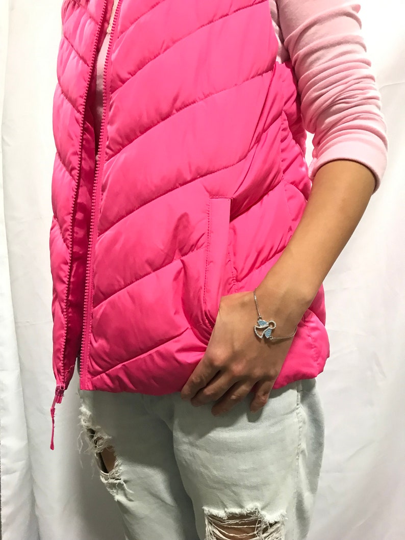 May include: A pink puffer vest with a zip fastening and a pocket on the left side. The vest is worn over a long-sleeved pink shirt and light blue jeans. The person is wearing a silver bracelet with a blue butterfly charm.