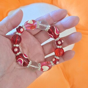 Puede incluir: Una pulsera de cuentas con cuentas rojas, rosas y transparentes. La pulsera presenta una mezcla de cuentas redondas y ovaladas con detalles plateados. Algunas cuentas tienen dise&ntilde;os de coraz&oacute;n y remolino blancos.