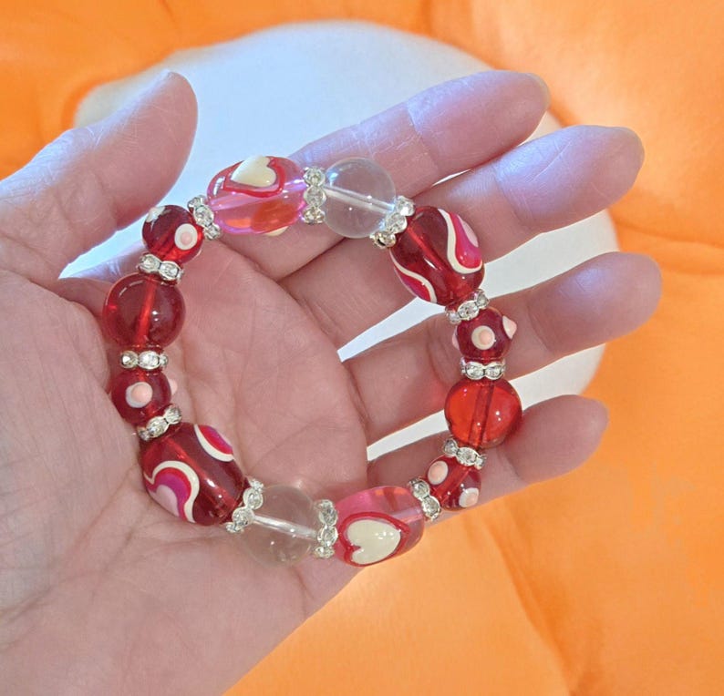 Puede incluir: Una pulsera de cuentas con cuentas rojas, rosas y transparentes. La pulsera presenta una mezcla de cuentas redondas y ovaladas con detalles plateados. Algunas cuentas tienen dise&ntilde;os de coraz&oacute;n y remolino blancos.