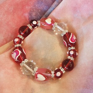 Puede incluir: Una pulsera de cuentas con cuentas de vidrio rojas, rosas y transparentes. Algunas cuentas tienen dise&ntilde;os de coraz&oacute;n blanco. La pulsera est&aacute; acentuada con espaciadores plateados y descansa sobre una tela rosa y morada.