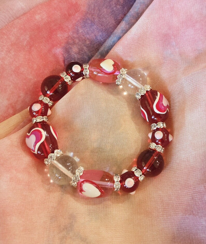 Puede incluir: Una pulsera de cuentas con cuentas de vidrio rojas, rosas y transparentes. Algunas cuentas tienen dise&ntilde;os de coraz&oacute;n blanco. La pulsera est&aacute; acentuada con espaciadores plateados y descansa sobre una tela rosa y morada.