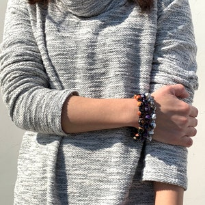 May include: A gray cowl neck sweater with a long sleeve and a textured pattern. The person is wearing a multi-colored beaded bracelet on their wrist.