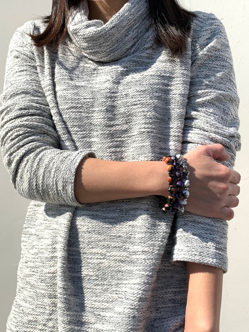 May include: A gray cowl neck sweater with a long sleeve and a textured pattern. The person is wearing a multi-colored beaded bracelet on their wrist.