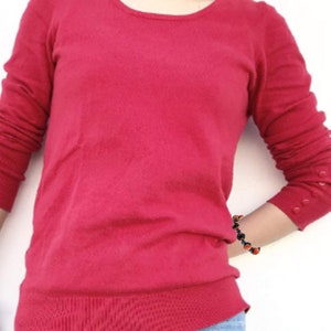 May include: A red long-sleeved sweater with a round neckline and button detailing on the cuffs. The sweater is worn by a person with a bracelet on their wrist.
