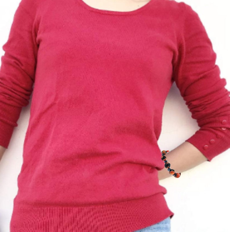 May include: A red long-sleeved sweater with a round neckline and button detailing on the cuffs. The sweater is worn by a person with a bracelet on their wrist.