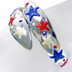 May include: A silver-toned bypass ring featuring a polished metallic finish decorated with small, colorful enamel stars in red, white, and blue. The design includes scattered dots and glittery accents on the star surfaces, creating a reflective appearance.