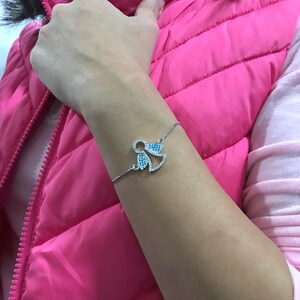 May include: A silver bracelet with a blue and white crystal angel charm. The bracelet is on a person's wrist.