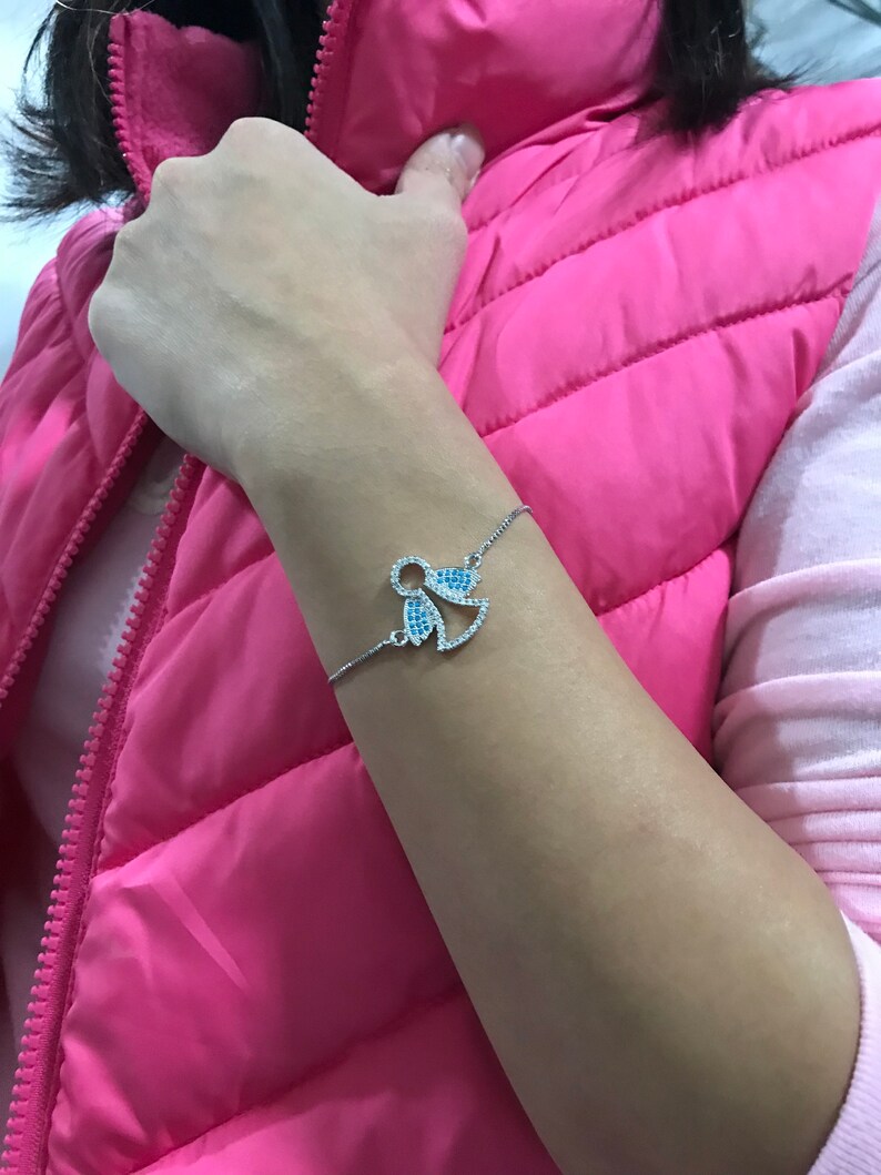 May include: A silver bracelet with a blue and white crystal angel charm. The bracelet is on a person's wrist.
