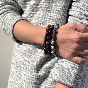 May include: Three stacked bracelets with colorful gemstone beads. The top bracelet has orange and black beads, the middle bracelet has purple and black beads, and the bottom bracelet has white and gray beads.