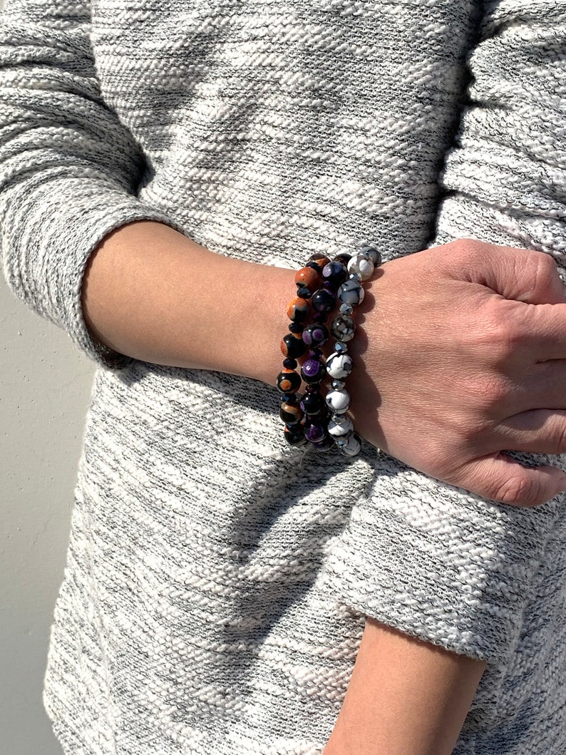 May include: Three stacked bracelets with colorful gemstone beads. The top bracelet has orange and black beads, the middle bracelet has purple and black beads, and the bottom bracelet has white and gray beads.