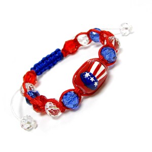 May include: A patriotic bracelet featuring red, white, and blue beads. The bracelet includes a central red bead with a design of a hat with stars and stripes. The bracelet is made with red cord and has a blue braided section.