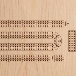 May include: Wooden cribbage board with a classic track design. The board features a series of holes for pegging, with a start and finish area. The board is made of light-colored wood, with the track and holes in a darker brown.