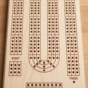 May include: A wooden cribbage board with a light wood finish. The board features three parallel tracks with evenly spaced holes for pegs. The word "START" is at the bottom left, and "FINISH" is at the bottom right.