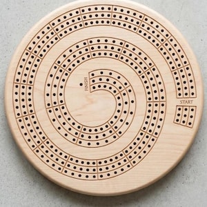 May include: A round wooden cribbage board with a spiral track design. The board is light-colored with numerous small holes for pegs and is labeled "START" and "FINISH". A classic game for family fun.