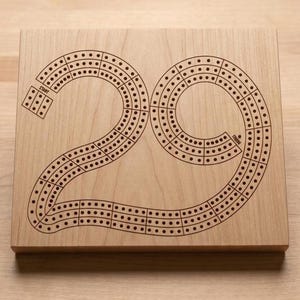 May include: A wooden cribbage board with a large "29" design etched into the surface. The board features a track of holes for pegging, with "START" and "FINISH" labels. The board is made of light-colored wood.