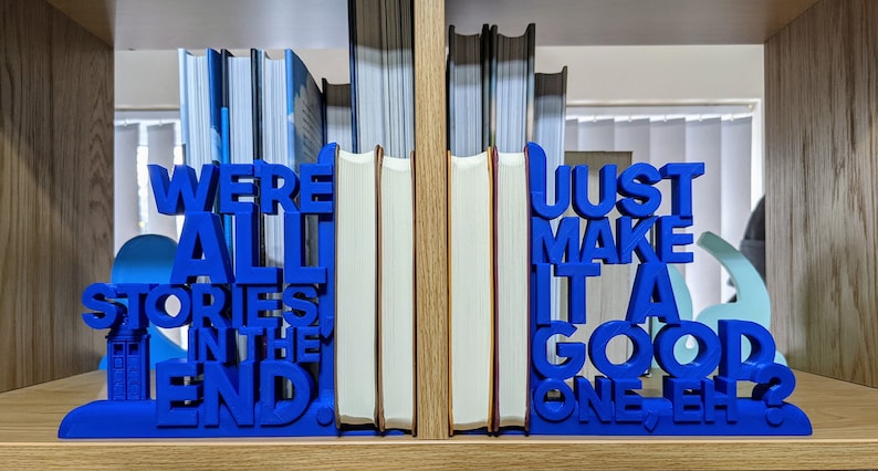 May include: A pair of blue bookends with the quote "We're all stories in the end. Just make it a good one, eh?" holding books on a wooden shelf. The bookends feature a blue police box design.