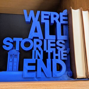 May include: A blue 3D-printed bookend with the quote "WE'RE ALL STORIES IN THE END." The bookend features a TARDIS design. The bookend is holding up several books with tan covers.