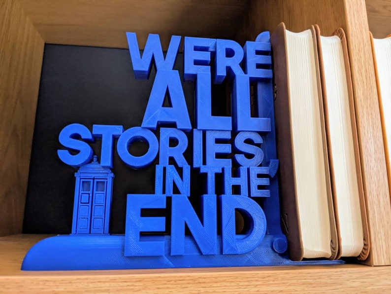 May include: A blue 3D-printed bookend with the quote "WE'RE ALL STORIES IN THE END." The bookend features a TARDIS design. The bookend is holding up several books with tan covers.