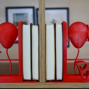 Könnte beinhalten: Rote Buchstützen mit Ballon und Text "You'll float too" und "Want a balloon?", die einen Stapel Bücher halten. Die Buchstützen bestehen aus einem festen Material und sind so konzipiert, dass sie Bücher auf einem Regal aufrecht halten.