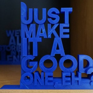 May include: A blue 3D-printed bookend, featuring the text "JUST MAKE IT A GOOD ONE, EH?". The bookend is positioned on a wooden shelf, with a second bookend and a book partially visible in the background.