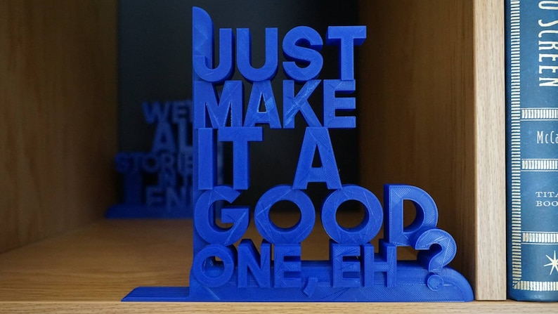May include: A blue 3D-printed bookend, featuring the text "JUST MAKE IT A GOOD ONE, EH?". The bookend is positioned on a wooden shelf, with a second bookend and a book partially visible in the background.