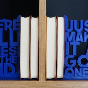 May include: Blue bookends with the quote "WE'RE ALL STORIES IN THE END" and "JUST MAKE IT A GOOD ONE-EH?". The bookends are holding several books with white pages and tan edges. The bookends have a TARDIS design.