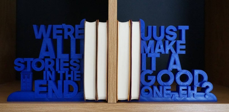 May include: Blue bookends with the quote "WE'RE ALL STORIES IN THE END" and "JUST MAKE IT A GOOD ONE-EH?". The bookends are holding several books with white pages and tan edges. The bookends have a TARDIS design.