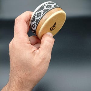 Può includere: Una piccola scatola di legno con una fascia a motivi bianco e nero attorno alla parte superiore. La scatola ha un coperchio di legno e una lettera "E" nera sul lato.