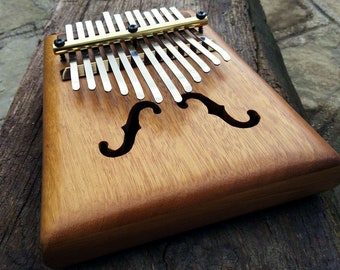May include: A wooden kalimba with a natural wood finish and a brass bar. The kalimba has 17 keys and a sound hole shaped like a violin.