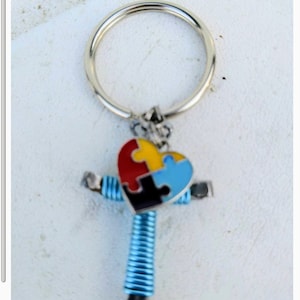 May include: A silver key chain with a blue, red, yellow, and black puzzle piece heart charm attached to a blue metal cross.