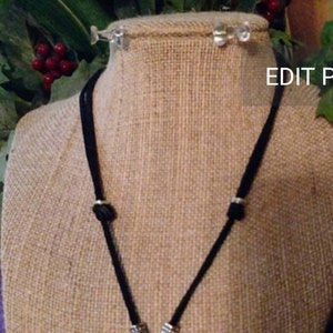 May include: A black cord necklace with silver accents on a burlap mannequin. The necklace is displayed against a green leafy background.