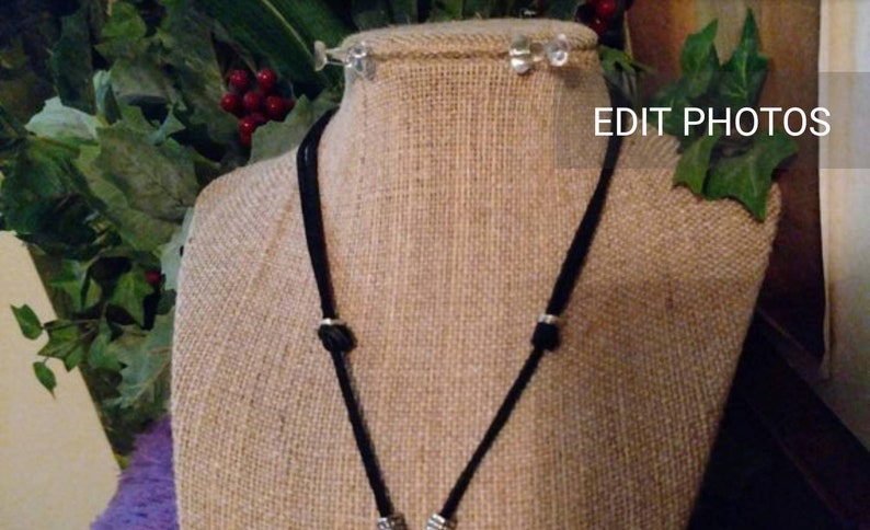 May include: A black cord necklace with silver accents on a burlap mannequin. The necklace is displayed against a green leafy background.