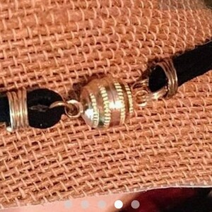 May include: A black suede cord bracelet with a gold-toned magnetic clasp. The clasp is round and has a textured design.