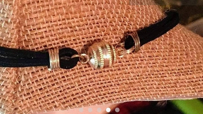 May include: A black suede cord bracelet with a gold-toned magnetic clasp. The clasp is round and has a textured design.