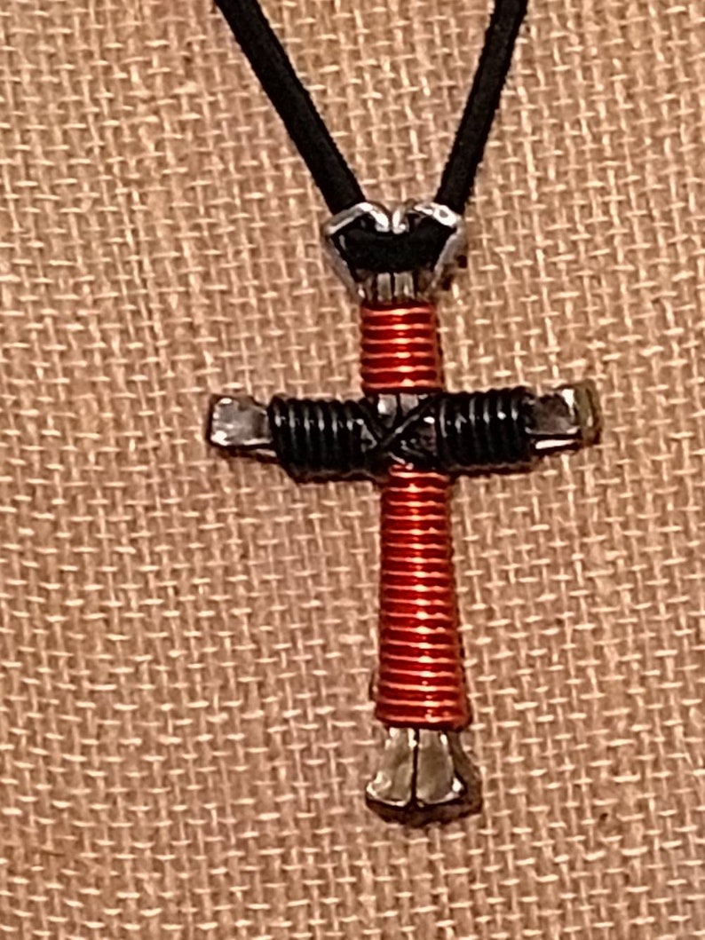 May include: A handcrafted cross pendant with a black cord. The cross is made from metal, wrapped with red and black wire. The cross is a religious symbol, and the pendant is suitable for everyday wear.