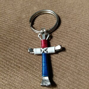 Puede incluir: Un llavero con forma de cruz de herradura rojo, blanco y azul. La cruz est&aacute; hecha de dos herraduras envueltas con alambre. El alambre es rojo, blanco y azul.