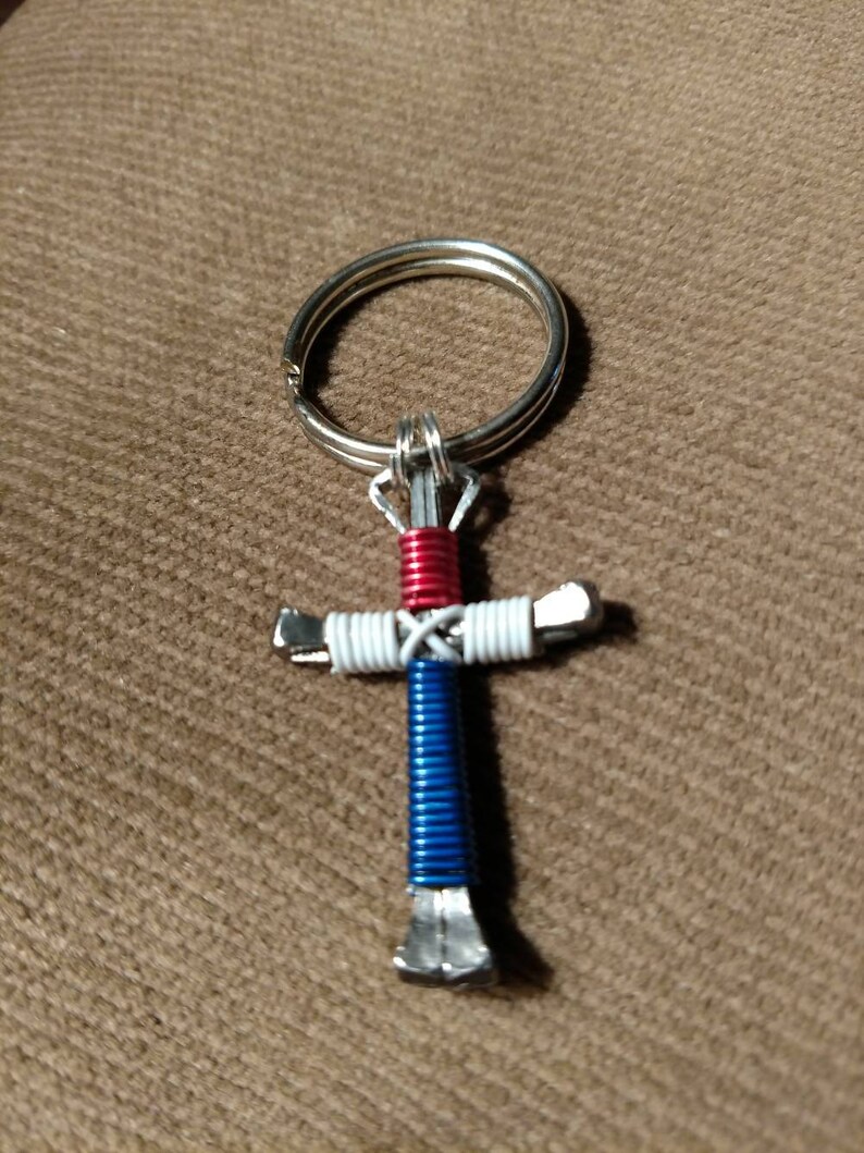 Puede incluir: Un llavero con forma de cruz de herradura rojo, blanco y azul. La cruz est&aacute; hecha de dos herraduras envueltas con alambre. El alambre es rojo, blanco y azul.