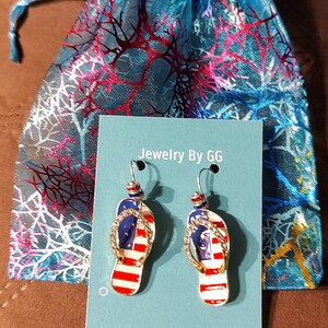 Puede incluir: Un par de pendientes de tono dorado con forma de chanclas con un dise&ntilde;o de bandera estadounidense. Los pendientes se muestran en una tarjeta azul con el texto "Jewelry By GG".