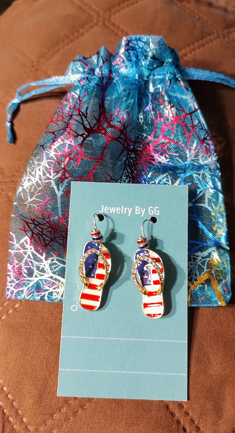 Puede incluir: Un par de pendientes de tono dorado con forma de chanclas con un dise&ntilde;o de bandera estadounidense. Los pendientes se muestran en una tarjeta azul con el texto "Jewelry By GG".
